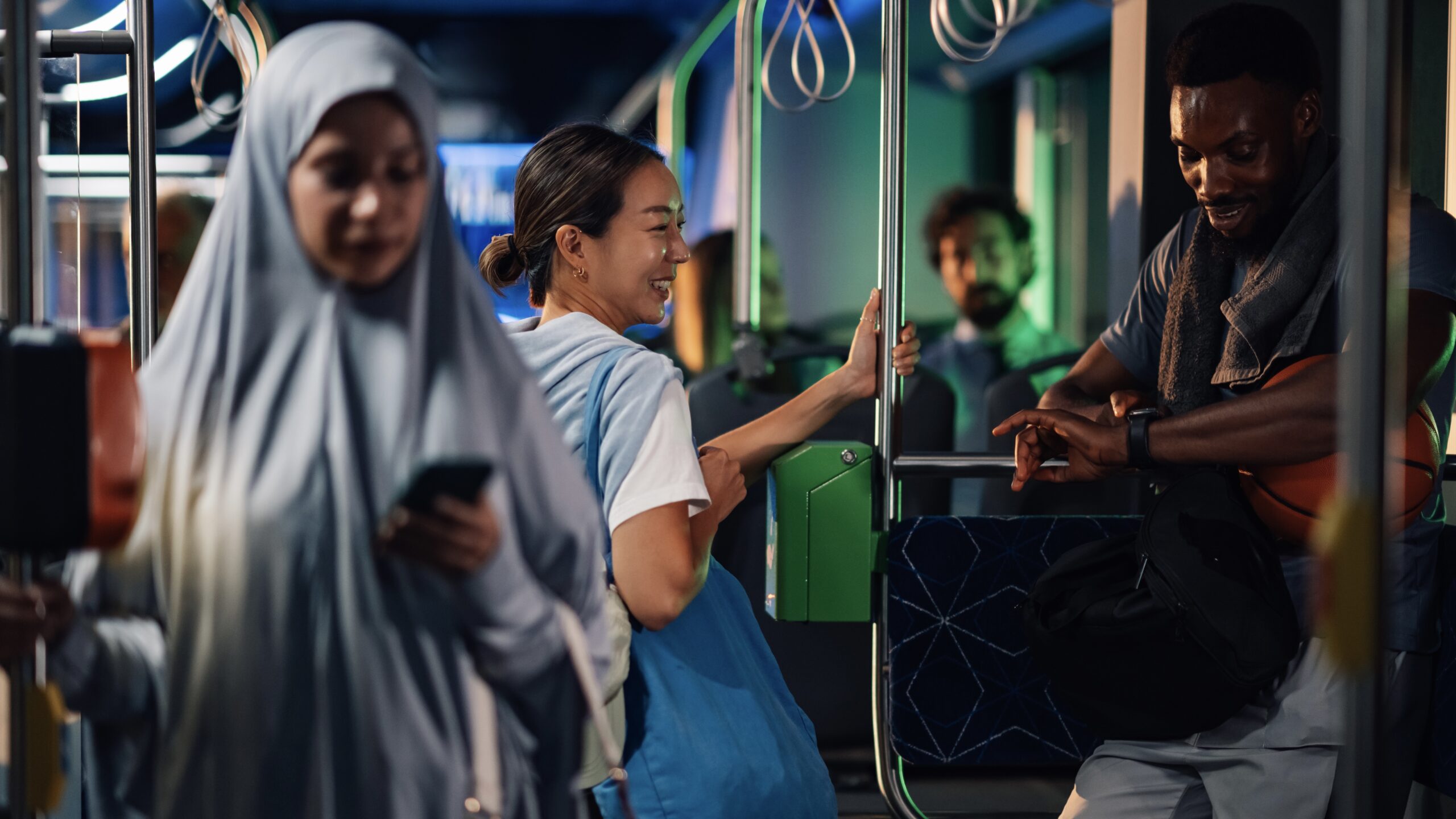 Passengers are using a modern public transport bus at night, holding onto the railing and looking at their phones and smartwatches